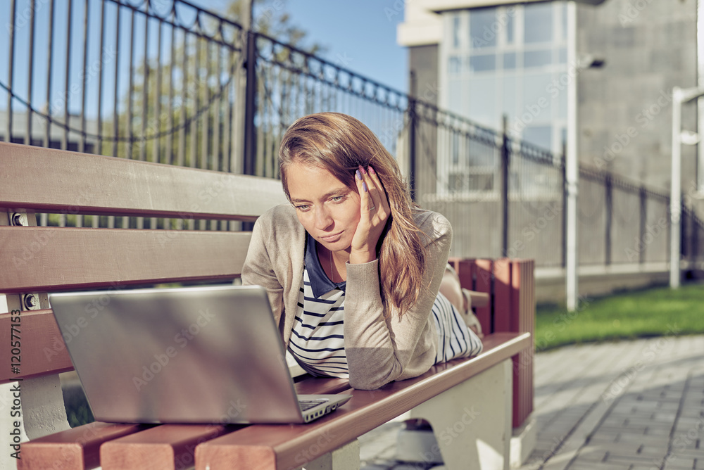 Fototapeta premium beautiful young brunette woman in a dress with blue and white stripes in sunglasses sitting on a bench and using laptop for surfing, chat, learn, game. Concept of urban social networking and internet.