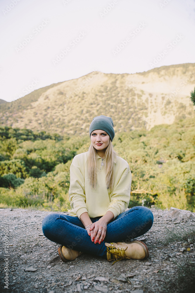 young blonde woman in a gray knitted hat, blue jeans and yellow working boots sitting outdoor on a background of mountains young blonde woman in a gray knitted hat, blue jeans and yellow working boots sitting outdoor on a background of mountains