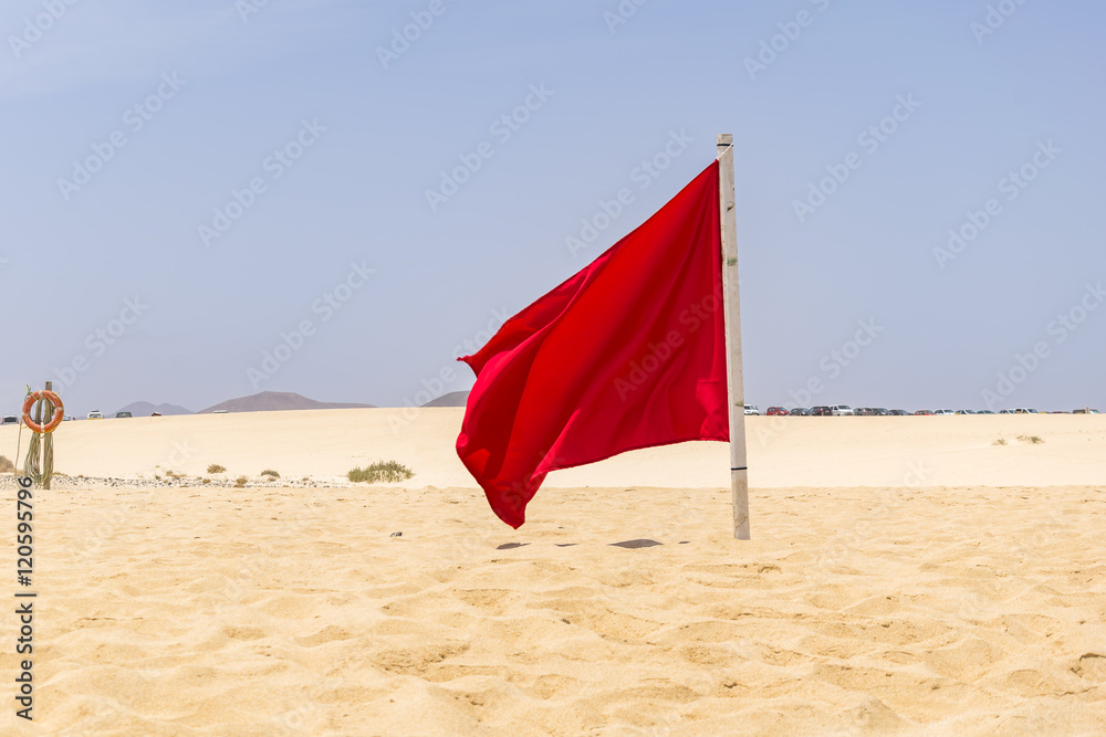 Red flag flying in the wind on the beach, photo taken in summer 2016 on Fuerteventura. Sunny day, as usual, no clouds on the sky and no people to see. Red flag flying in the wind on the beach, photo taken in summer 2016 on Fuerteventura. Sunny day, as usual, no clouds on the sky and no people to see.