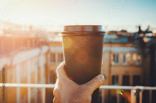 hands holding hot craft cup of coffee or tea in morning sunlight with view to blurred city background. Enjoy, lifestyle, take away breakfast concept. woman on the roof with hot drink
