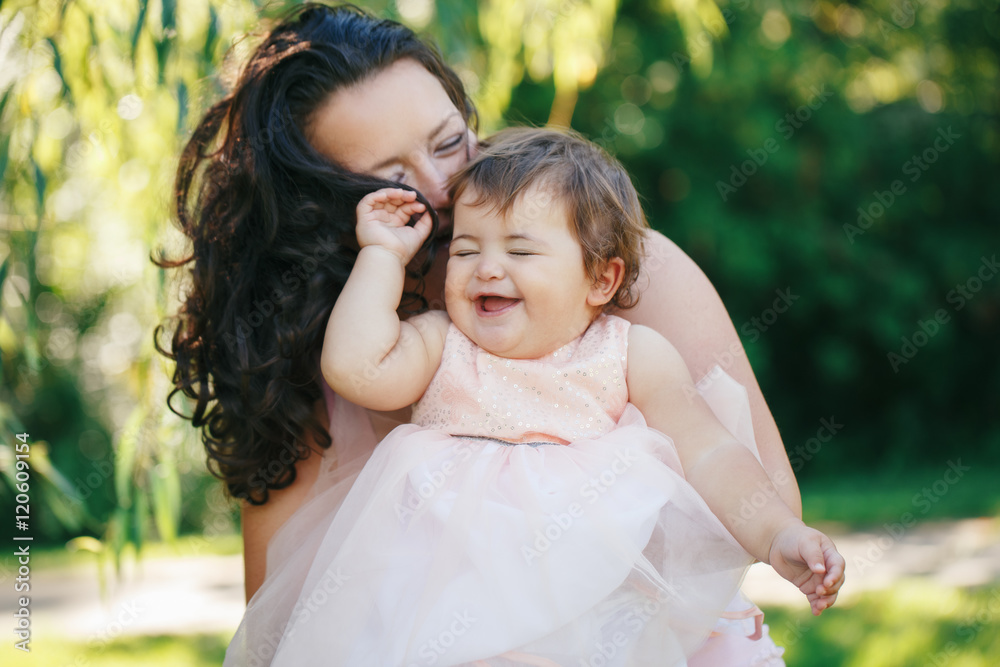 Closeup group portrait of beautiful white Caucasian brunette mother holding laughing baby daughter kissing her in cheek on sunny spring summer day in park outside