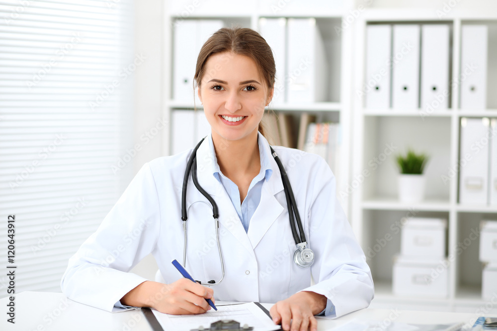 Young brunette female doctor sitting at the table and working at hospital office. Health care, insurance and help concept. Physician ready to examine patient Young brunette female doctor sitting at the table and working at hospital office. Health care, insurance and help concept. Physician ready to examine patient