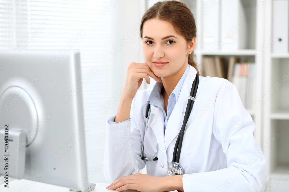 Young brunette female doctor sitting at the table and working at hospital office. Health care, insurance and help concept. Physician ready to examine patient Young brunette female doctor sitting at the table and working at hospital office. Health care, insurance and help concept. Physician ready to examine patient
