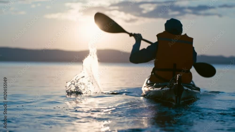 Slow motion rear view of male kayaker paddling away from camera towards sun setting above chain of mountains 