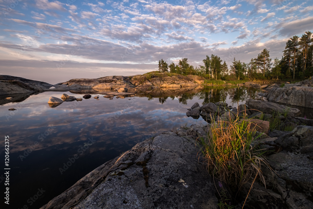 A bush of grass lighten by the evening sun is growing on granite rocks. In the background clouds reflected in smooth water surface of a small bay, Ladoga, Karelia