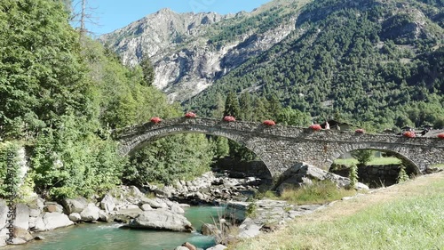 Gaby, Aosta valley, Italy. People walking on old medieval bridge with mountains landscape, Lys river and flowers on the ancient path. Summer holidays, nature and italian tourism