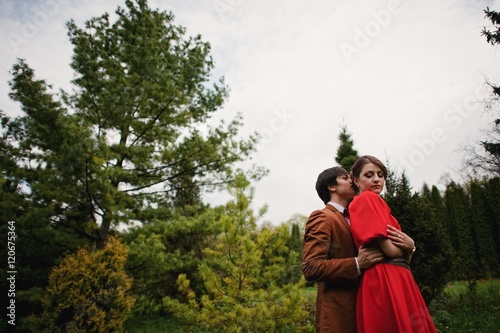 Couple hugging in love at beautiful landscape of trees and meadows. Stylish man at velvet jacket and girl in red dress in love together