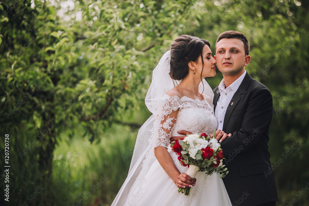 couple in wedding attire with a bouquet of flowers and greenery is in the hands against the backdrop of the garden at sunset, the bride and groom couple in wedding attire with a bouquet of flowers and greenery is in the hands against the backdrop of the garden at sunset, the bride and groom
