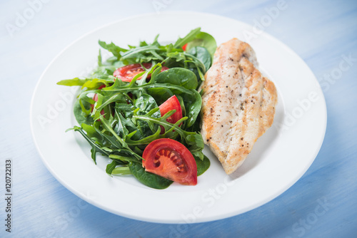 Roasted chicken breast and fresh salad with tomato and greens (spinach, arugula) close up on blue wooden background. Healthy food.
