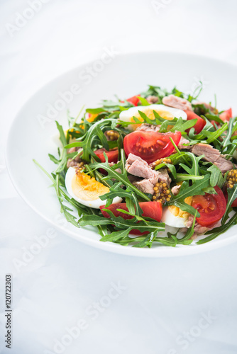 Fresh salad with tuna, tomatoes, eggs, arugula and mustard on white textured background close up. Healthy food.