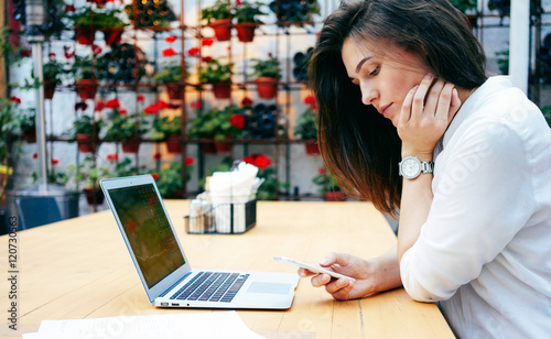 A charming brunette student girl is sitting  in a modern coffee shop and using digital cell phone. A beautiful businesswoman with long brown hair is using a mobile phone while sitting with a laptop.