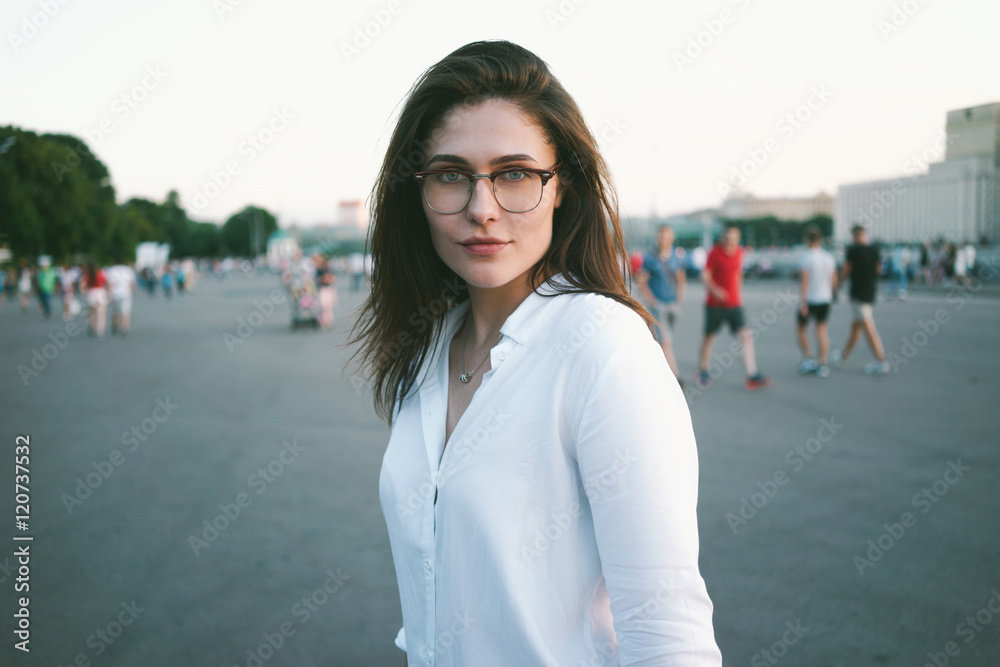 A waist up photo of a beautiful brunette woman wearing white shirt. A charming girl with long brown hairs wearing glasses is looking at the camera while standing on a city park background.