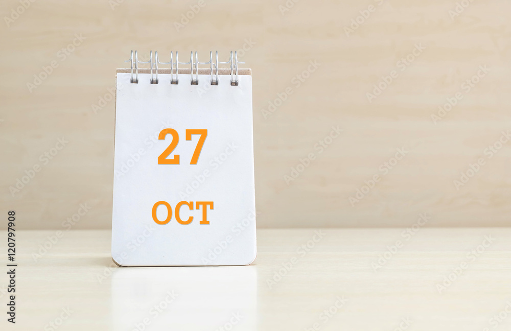 Closeup surface note book with orange 27 oct word in page on blurred brown wood desk and wood wall textured background with copy space under window light Closeup surface note book with orange 27 oct word in page on blurred brown wood desk and wood wall textured background with copy space under window light