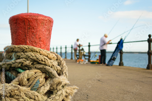 Red stone mooring bollard with rope tied around it on Torquay pier where unidentified fishermen prepare their fishing lines
 photo