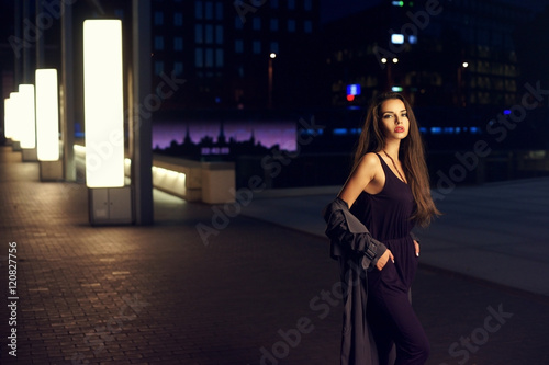 Fashion portrait of young beautiful pretty woman with long curly hair standing in night city and looking at you