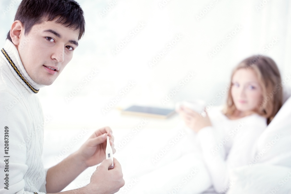 Young man sitting with thermometer with his wife in the background and looking at camera Young man sitting with thermometer with his wife in the background and looking at camera