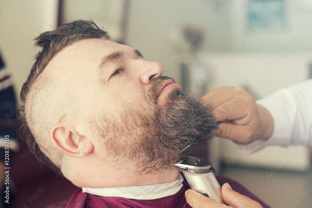 Fototapeta premium Male barber cuts the beard using clipper of a adult man with a mohawk