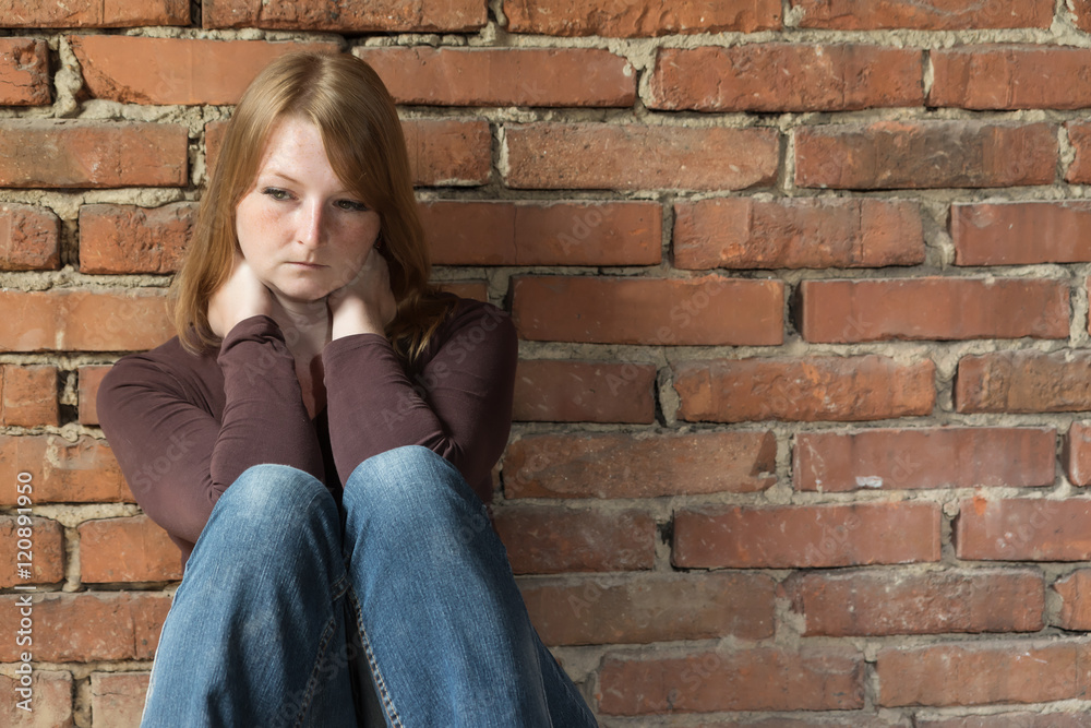 Sad redhead young woman is sitting in natural light in front of an old brick wall.