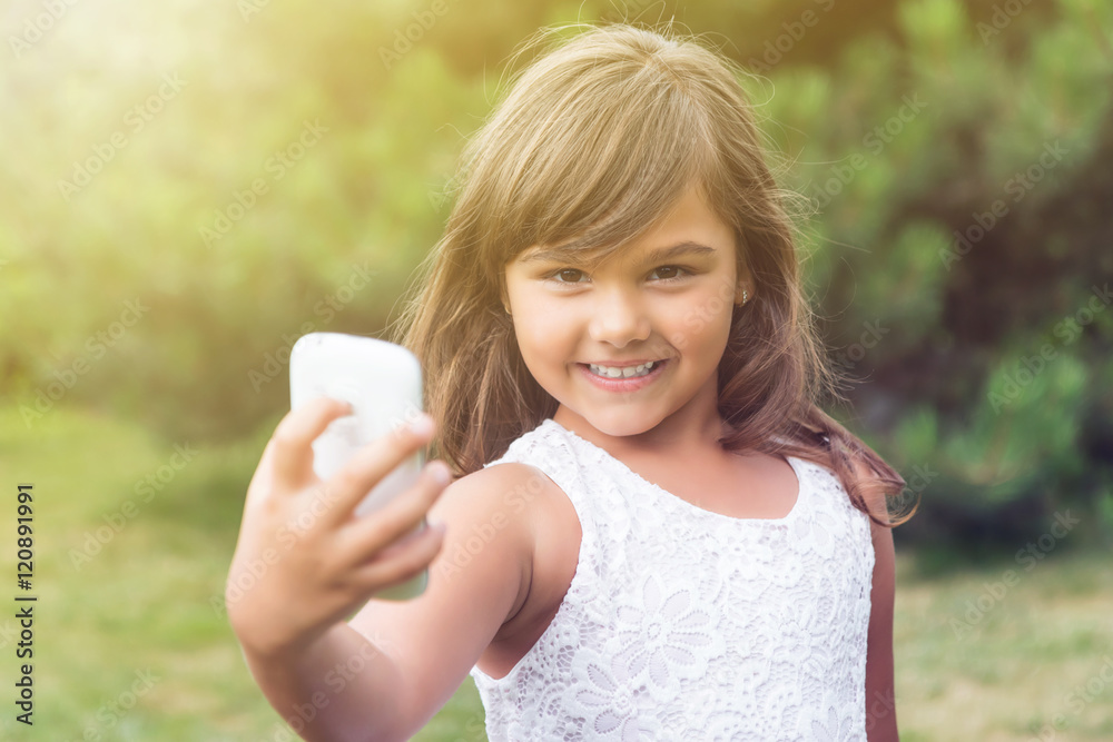 Smiling attractive little girl is doing selfie in summer light. Girl is looking at the camera holding smart phone in her hand. All potential trademarks are removed. Smiling attractive little girl is doing selfie in summer light. Girl is looking at the camera holding smart phone in her hand. All potential trademarks are removed.