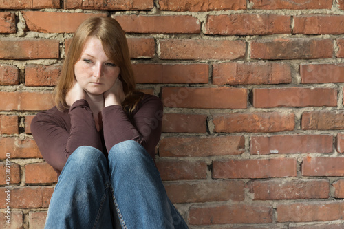 Sad redhead young woman is sitting in natural light in front of an old brick wall. © frank11