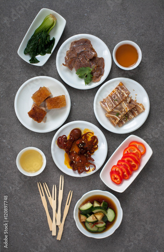 Assorted Asian appetizers on white plates: octopus in spicy sauce, sliced beef shank, pigs ears, crispy barbecue pork . Dark gray background 