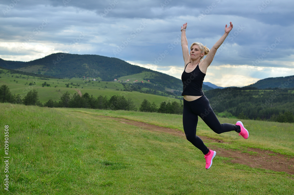 Joyful sportswoman in pink sneakers jumping on a meadow with hills and cloudy sky in background Joyful sportswoman in pink sneakers jumping on a meadow with hills and cloudy sky in background