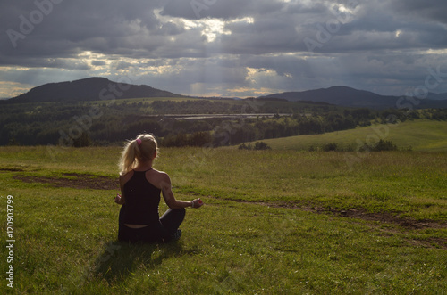 Woman who is meditating in a mountain meadow in late afternoon with face turned to the sun behind clouds on a stormy sky
