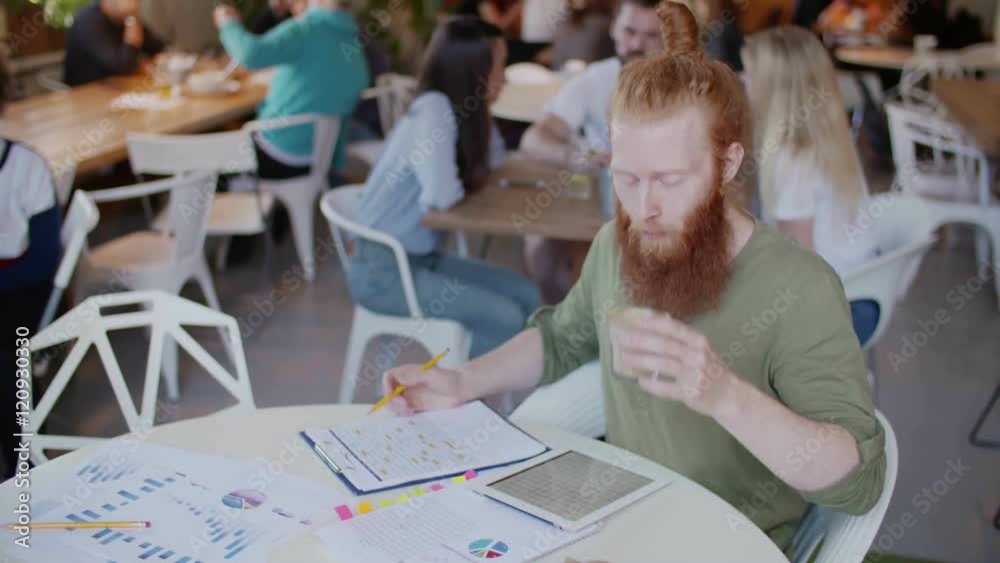Ginger man working in cafe and drink coffee on table graph and papper. Behind him cheerful group of young peoples