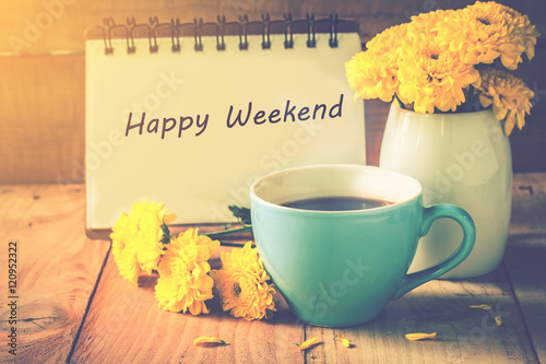blue cup of coffee on wooden floor with yellow flower in white pot and happy weekend note on morning sunlight. vintage color tone, happy weekend concept.
