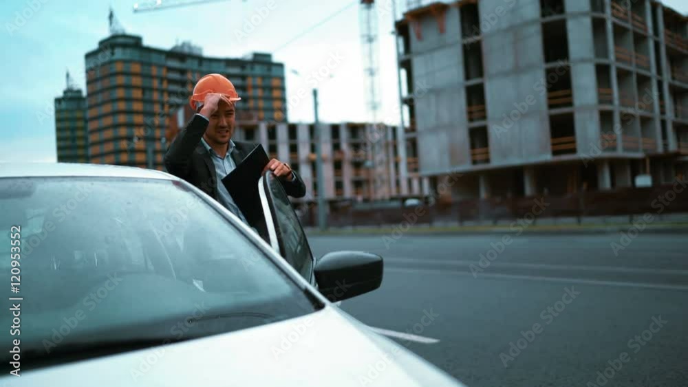 4k. Ultra HD. Young successful architect businesman exit his white car wear the orange helmet and walk to newly constructed building object with crane and beams. Textpad with drawing and notes 4k. Ultra HD. Young successful architect businesman exit his white car wear the orange helmet and walk to newly constructed building object with crane and beams. Textpad with drawing and notes