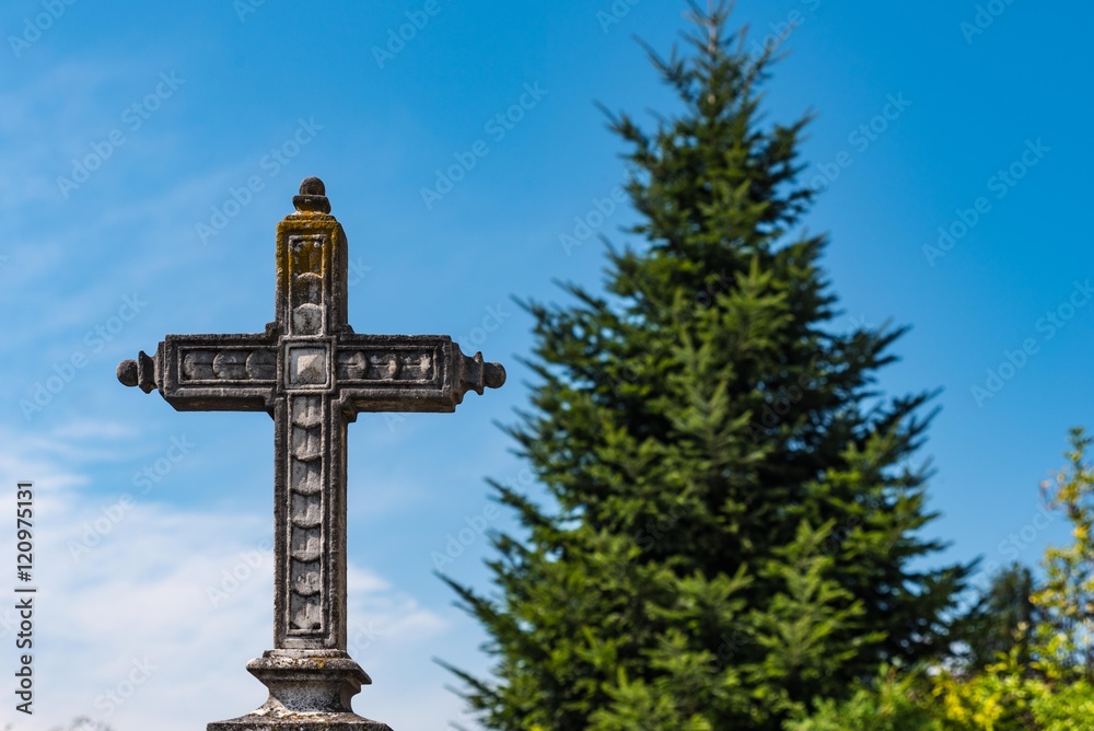 Steinernes Kreuz am Friedhof vor blauem Himmel und großem Fichtenbaum