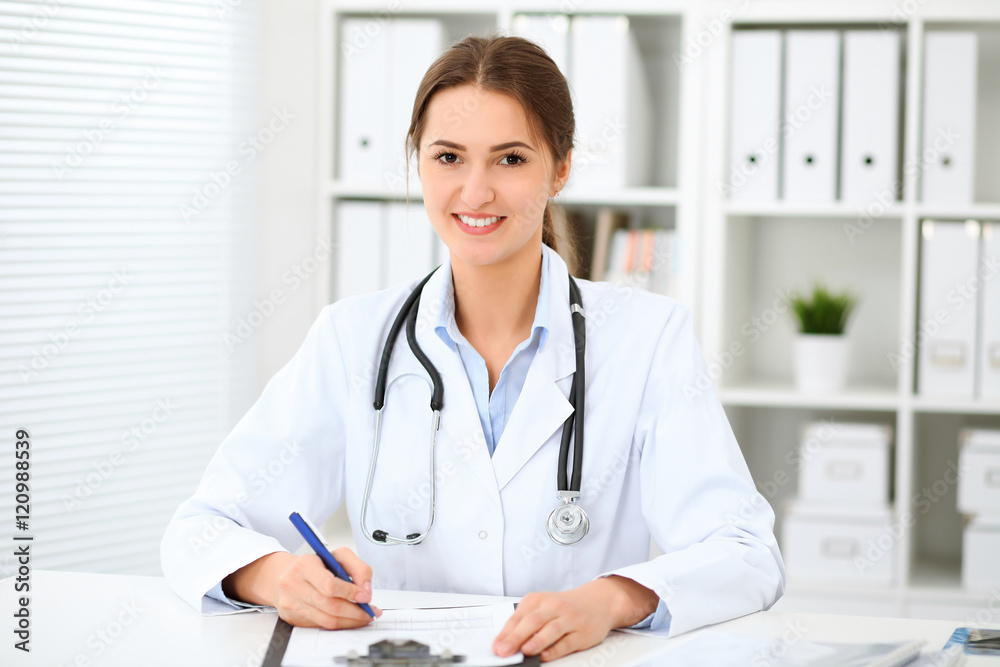 Young brunette female doctor sitting at the table and working at hospital office. Health care, insurance and help concept. Physician ready to examine patient Young brunette female doctor sitting at the table and working at hospital office. Health care, insurance and help concept. Physician ready to examine patient