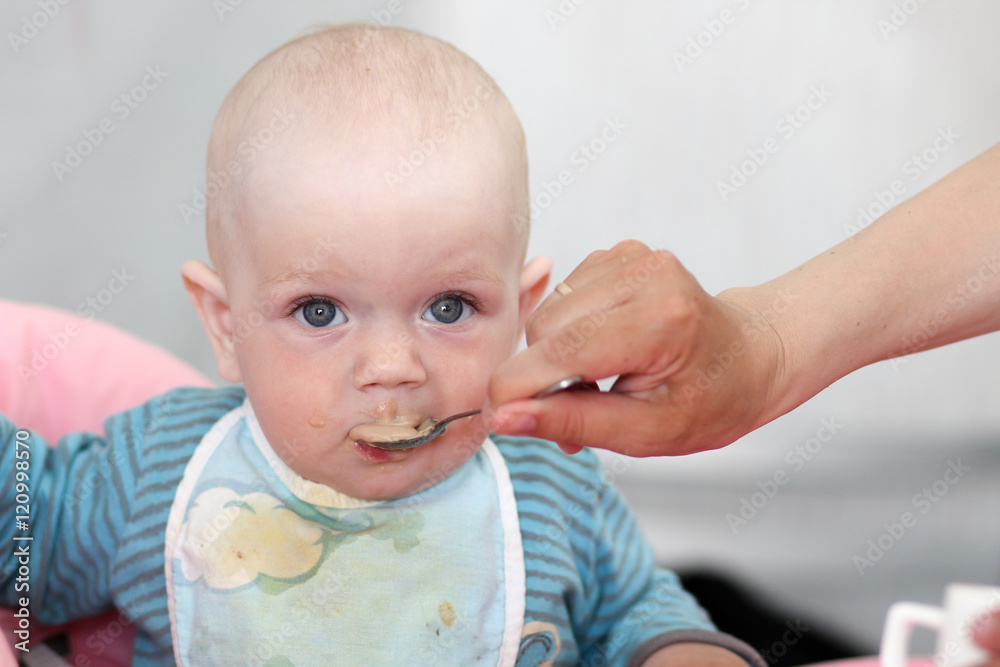 Beautiful baby eats porridge from mom's hand. He is sitting on a pink children' chair.