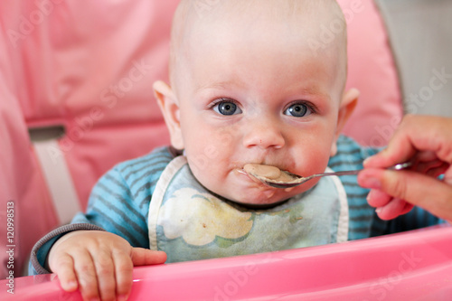 Beautiful baby eats porridge from mom's hand. He is sitting on a pink children' chair.