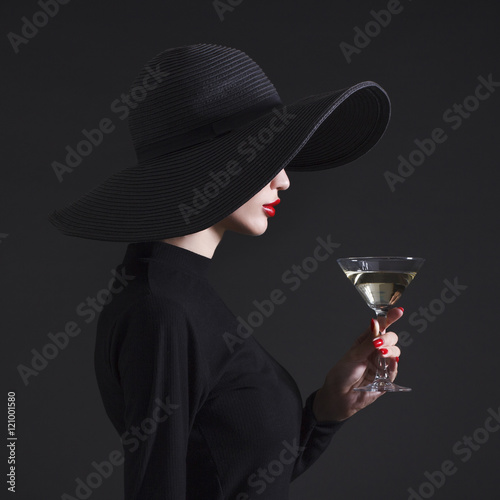 Luxury woman in a large black hat and bright lips with a glass of Martini.