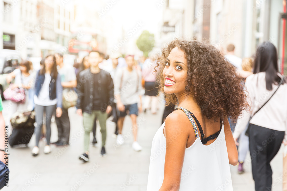 Young woman looking at camera while walking in London. Mixed race girl looking back and smiling. Blurred people on background walking on the sidewalk - obrazy, fototapety, plakaty 