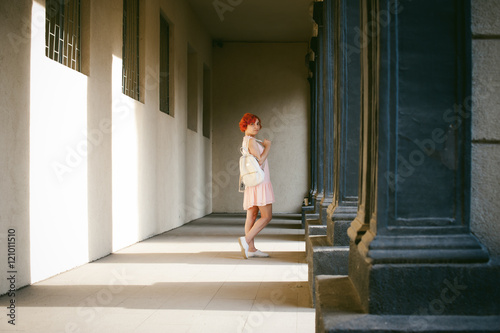 girl in a pale pink dress with dyed red hair, a white backpack in hand, walking between light and shadow on the columns sunny summer day © evgeniykleymenov
