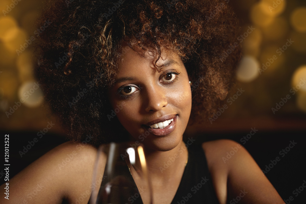 Young attractive African woman with happy cheerful smile, holding glass of beverage or alcohol drink, spending New Year's eve at fashionable restaurant. Joyful African woman relaxing at nightclub