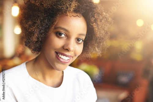 Portrait of beautiful African woman with curly hair, dressed in casual top, looking and smiling at camera, showing her white teeth. Black girl relaxing at coffee shop, waiting for her boyfriend