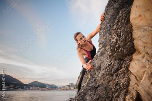 Portrait of young female rock climber overhanging cliff. Beautiful blue sky, city mountains on the background. © fesenko