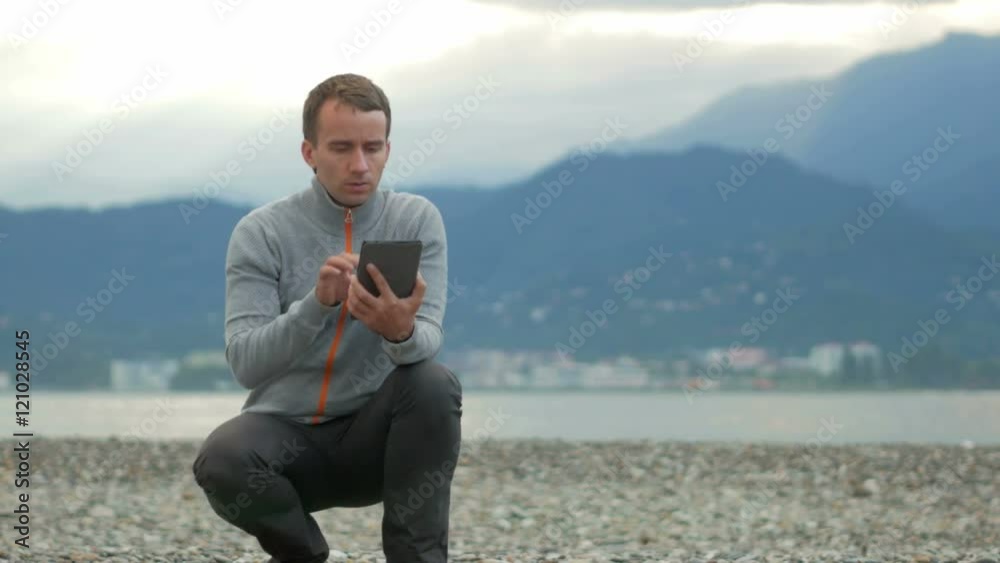 Young handsome man sitting on the rocky beach. He checks messages on the tablet. Behind the Pacific Ocean and the mountains.