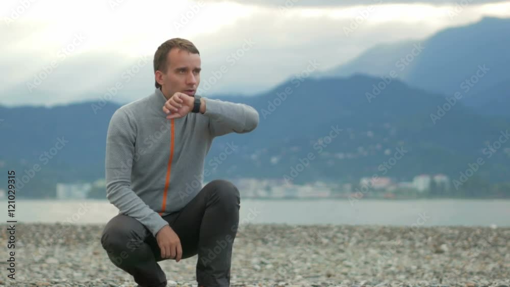 Young handsome man sitting on the rocky beach. He checks messages on the smart watch. Behind the Pacific Ocean and the mountains. Young handsome man sitting on the rocky beach. He checks messages on the smart watch. Behind the Pacific Ocean and the mountains.