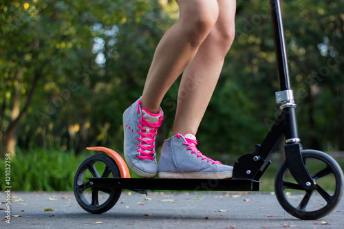 Female rider in gray shoes with pink laces is riding on a black kick scooter on asphalt road in a park with landscape on a background. Close-up horizontal view photo