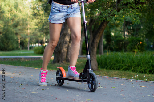 Athlete female rider in gray shoes with pink laces is riding on a black kick scooter on asphalt road in a park. Close-up horizontal view