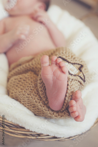 Closeup of legs of a newborn baby peeking out of light brown knitted blankets,which the baby covered from the waist up,the baby sleeps on a white soft blanket in a large wicker basket
