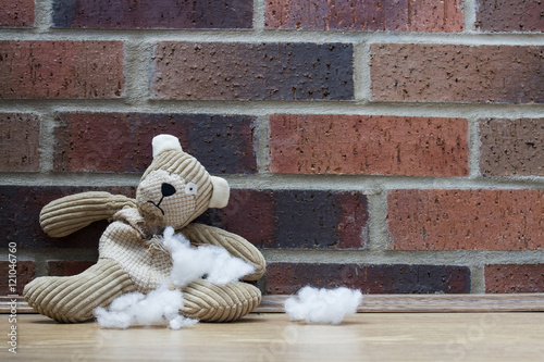 A sad, abandoned teddy bear with stuffing and filling falling from a hole in his ripped and torn tummy and sitting alone against a brick wall.