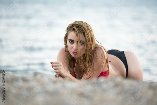 Blond woman with desired curved body wear two pieces bikini lying prone on rocky sand, sea as background photo