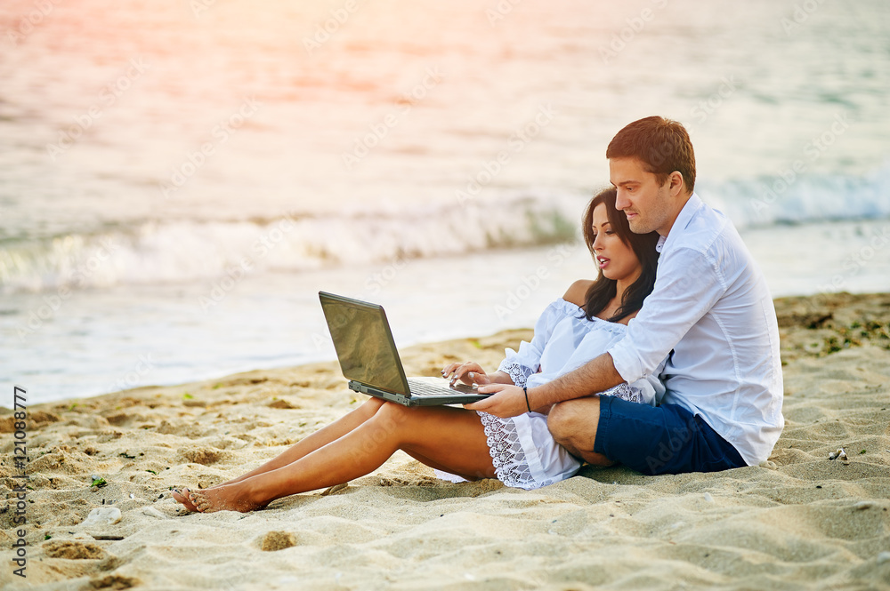 Beautiful romantic couple relaxing on the beach . By browsing on the laptop