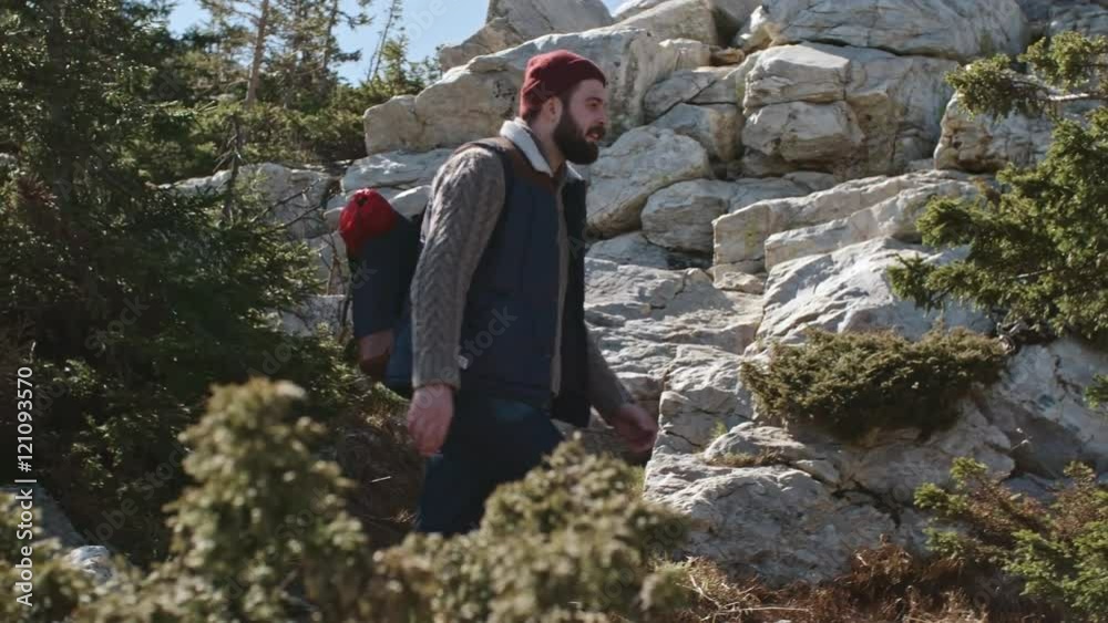 Slow motion tracking of bearded young man with backpack walking along trail high up in mountains