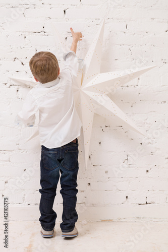 Back view on little toddler boy decoration wall with Christmas star. Child putting decorations on the walls for holidays season.